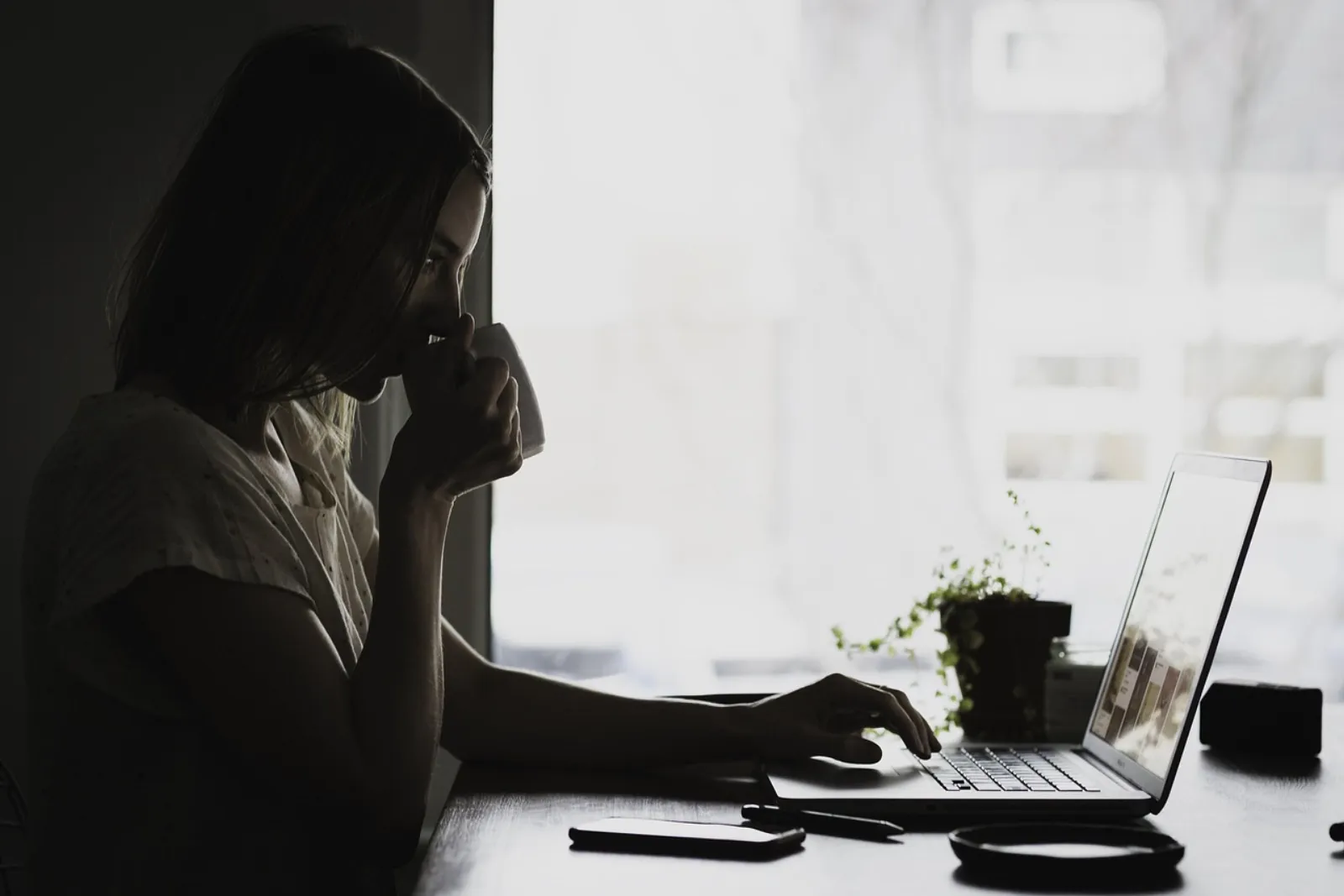 Person som läser på en laptop vid ett fönster med en kaffekopp i handen